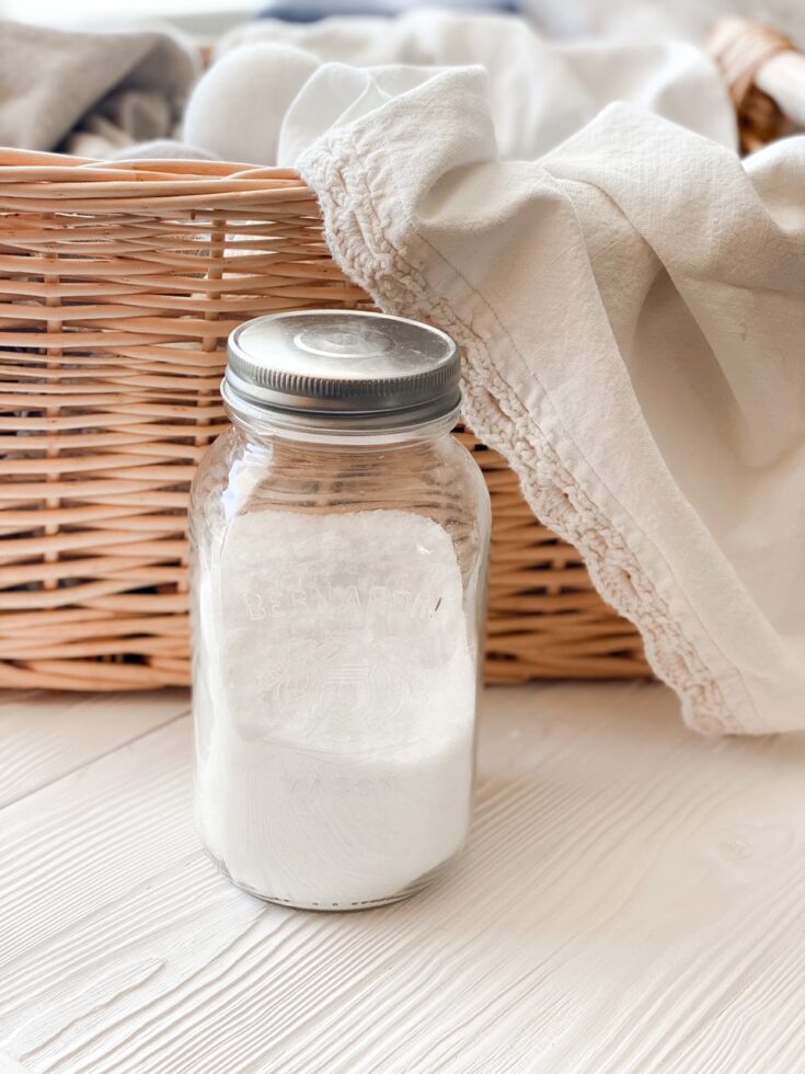 mason jar of homemade fabric softener on white wood backdrop in front of wicker basket filled with dish towels