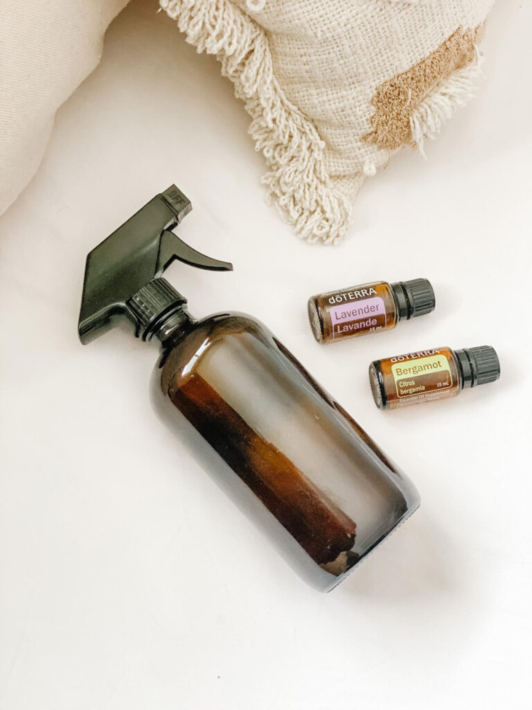 amber glass spray bottle laying on bed with linen spray beside lavender and bergamot essential oil