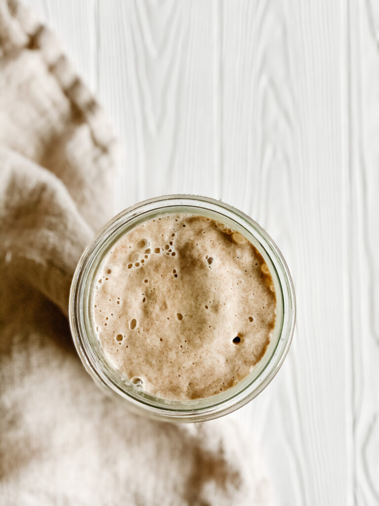 overhead shot of active gluten free sourdough starter in a glass jar on a white wood backdrop with a beige dish towel