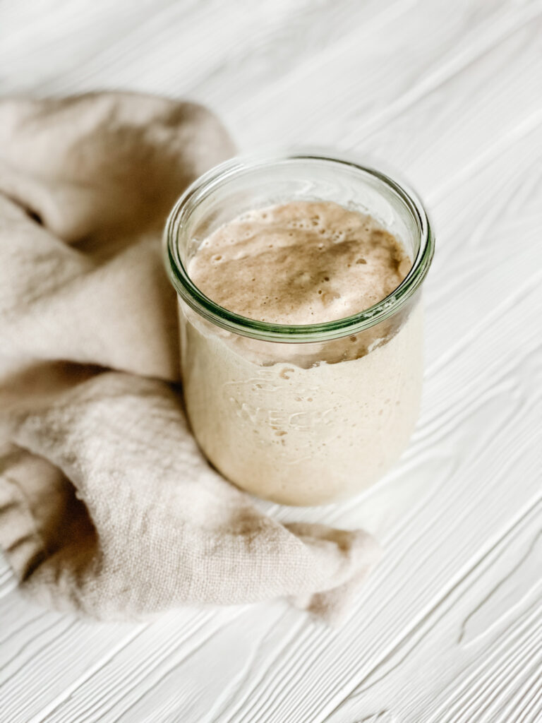 a jar of bubbly gluten free sourdough starter on a white wood backdrop with a beige dish towel