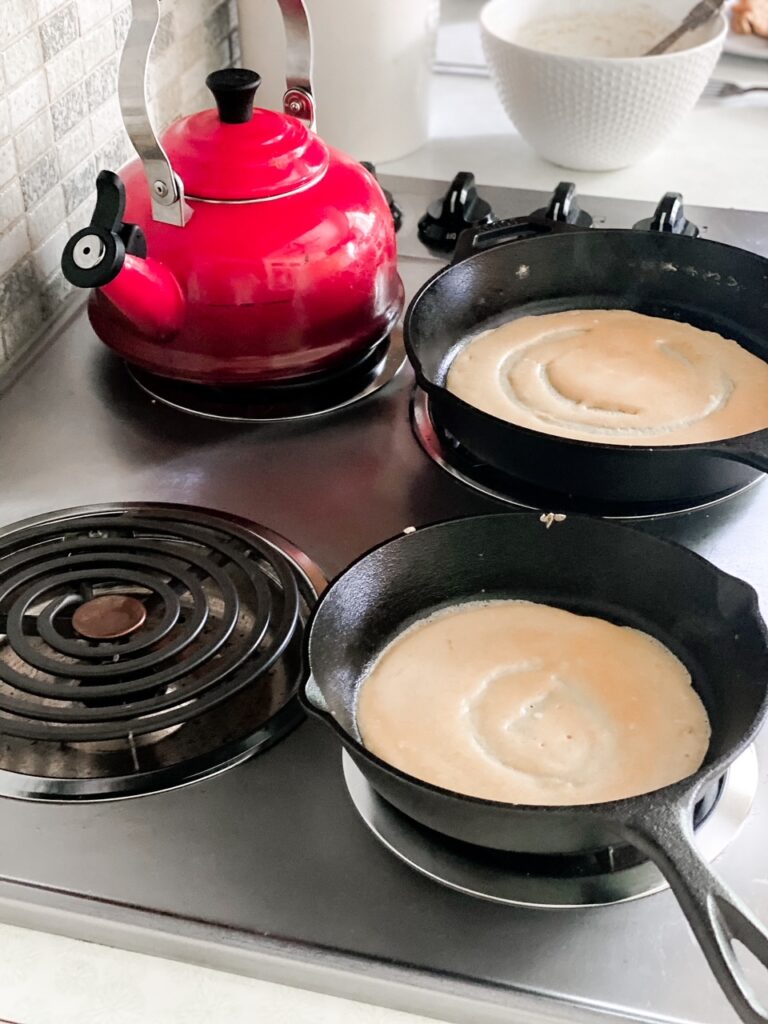 two cast iron pans with sourdough crepes cooking and red kettle and white bowl in the background
