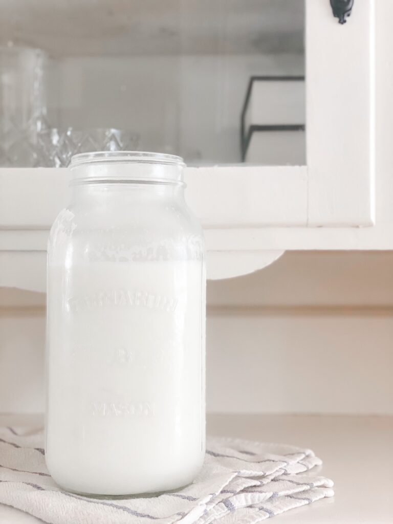 half gallon jar of raw milk on a white and blue dish cloth in front of a cream china cabinet