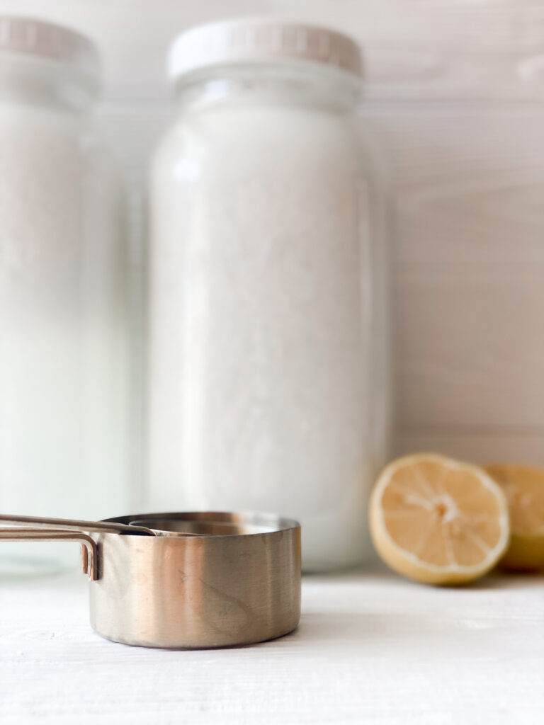 two half gallon jars of laundry soap with gold measuring cups and a lemon sliced in half