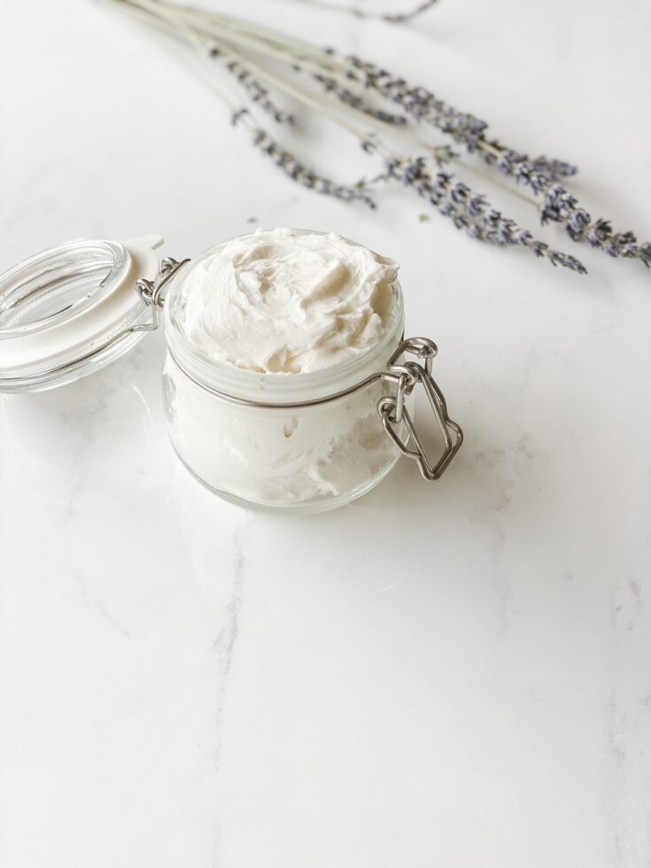 glass jar filled with homemade natural body butter on marble surface and lavender in the background