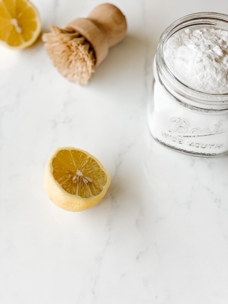 marble background with a sliced lemon, wood scrub brush, and jar of baking soda
