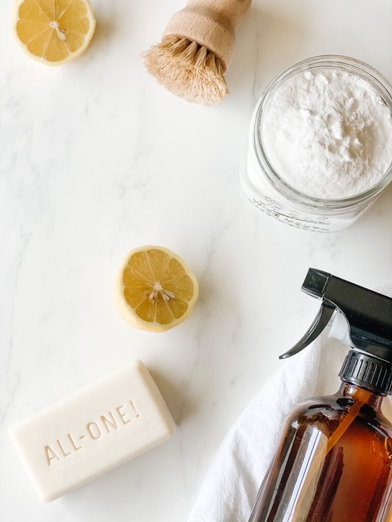 marble back drop with a sliced lemon, castile soap, amber spray bottle, wood scrub brush and baking soda